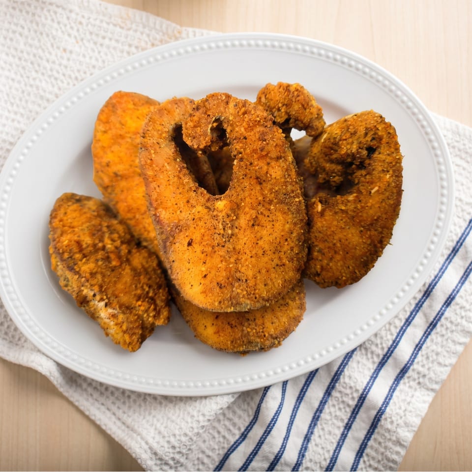 A white platter filled with golden-brown fried fish pieces, seasoned with dark spices, on a white and blue striped towel.